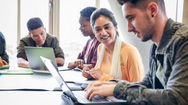 Young woman smiles and looks over at her peers laptop.