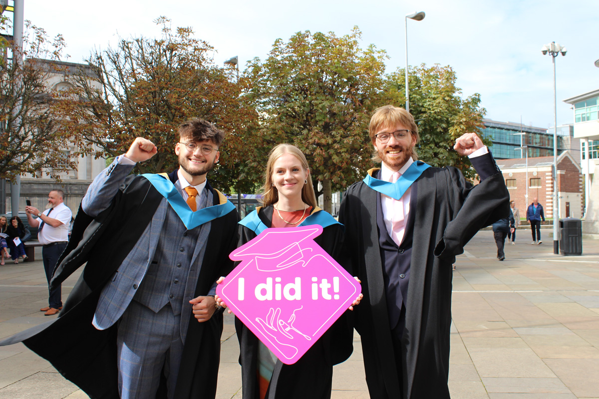 Three students in graduation gowns with an I did it sign.