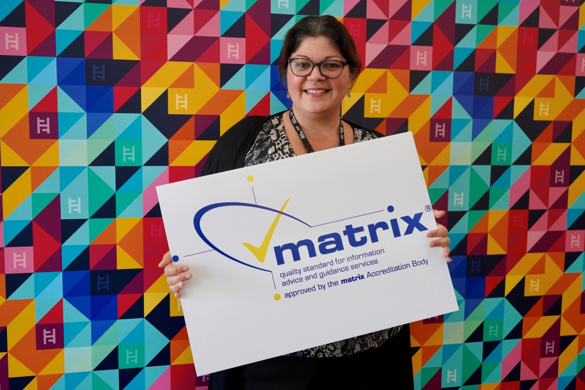 Head of Learner Services at Hull College, Shelley Newton, standing in the reception area of the Queen's Gardens campus, holding the matrix Standard logo. The college's modern interior is visible in the background, emphasising a welcoming and professional environment."