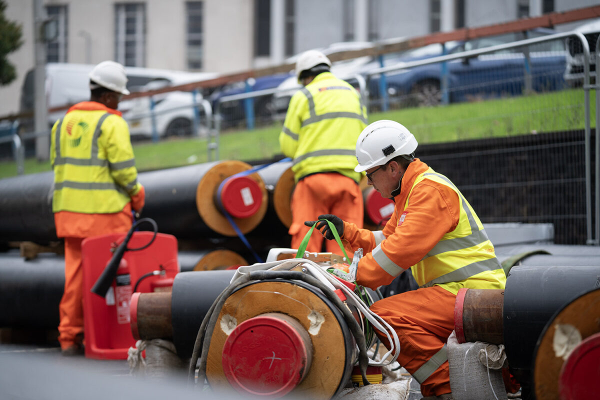 Three engineers wearing high-vis jackets and hard hats work on installing large pipes outside.