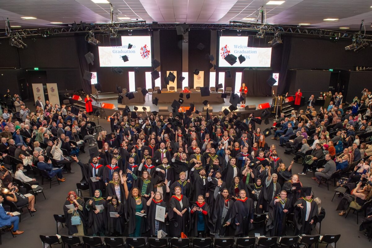 An aerial shot where a hall full of graduates throw their graduation caps into the air with the stage in the background.