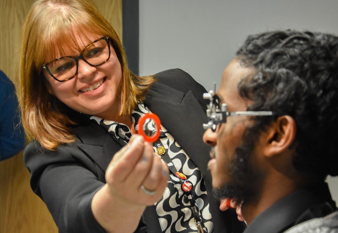 A female ophthalmic lecturer shows how to test vision using optical equipment on a male student who is sat in a mock optician's chair at college.