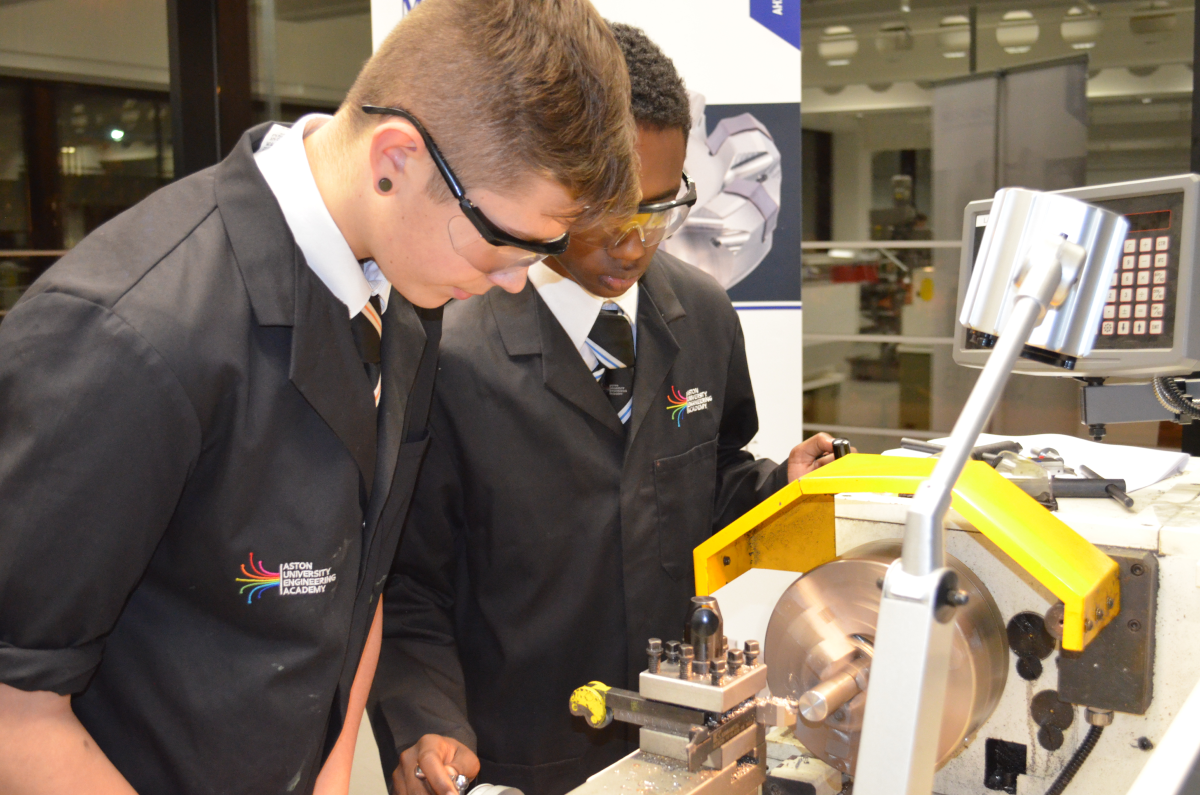 Two students in lab coats and safety glasses operate a lathe machine in a workshop setting.