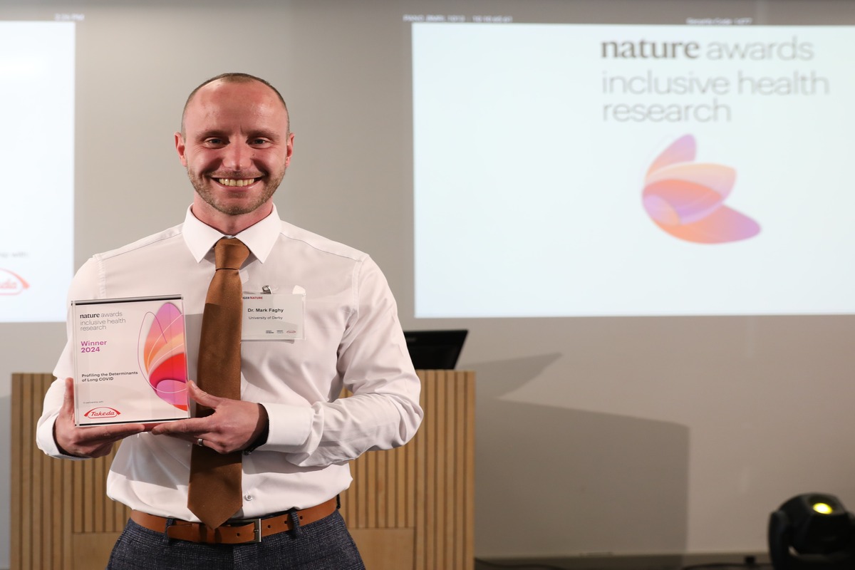 A smiling man stands holding a certificate after winning an award for research into long covid