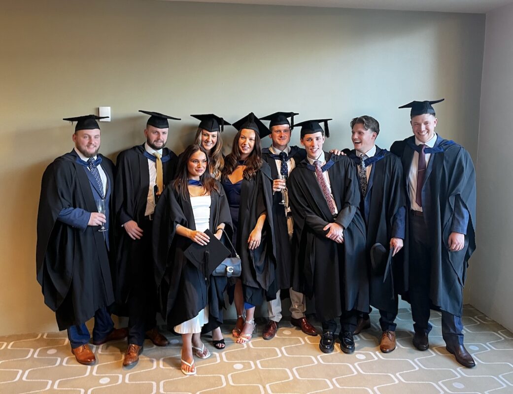 A mixed group of nine Leeds College of Building students pose together in a function room in their graduation cap and gowns.