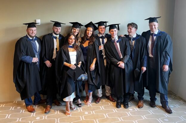 A mixed group of nine Leeds College of Building students pose together in a function room in their graduation cap and gowns.