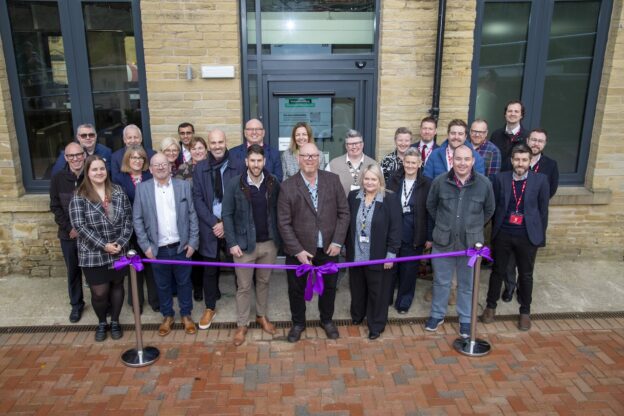 Bradford College Principal & CEO Chris Webb stands in the centre of a group of gathered guests to mark the opening of Garden Mills. Chris holds a pair of scissors and prepares to cut a purple ribbon. The group poses in front of the main entrance to the stone building.