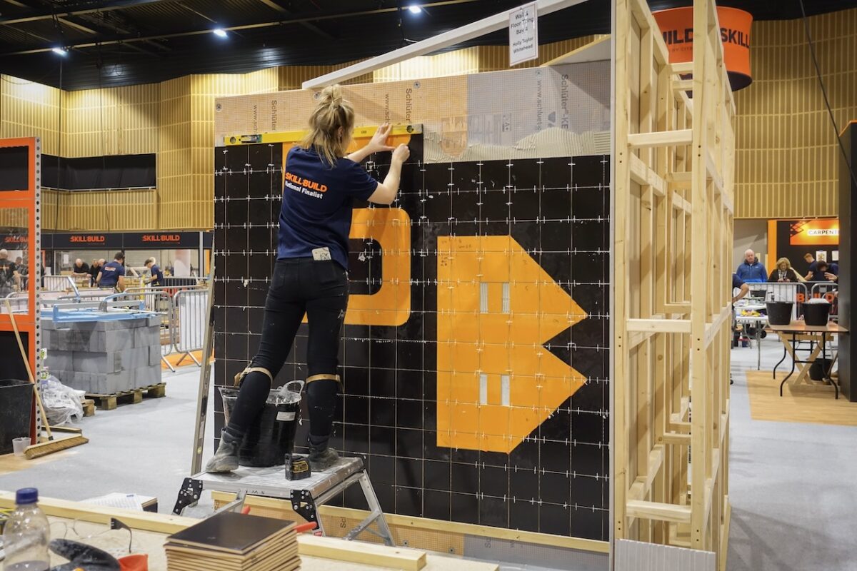 A female student stands on a step ladder completing a tiling project wall that spells out the initials SB in orange and black. The student is in a construction competition warehouse.