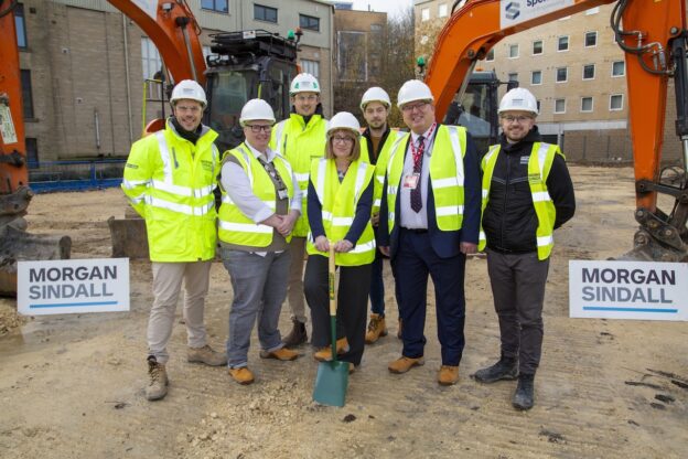 A group of executives wearing high-vis jackets, hard hats and boots, stand together on a building site with diggers in the background. The central female figure is posing holding a spade.