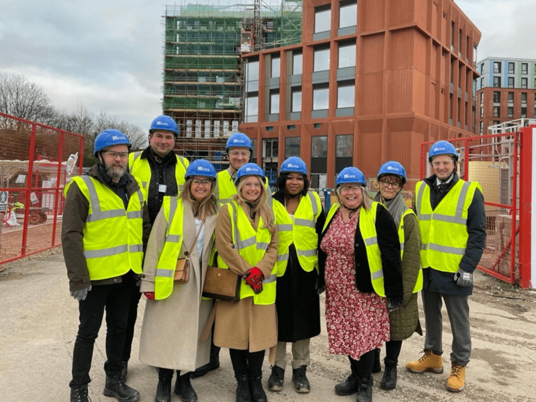 People wearing high vis and hard hats are standing in a row, smiling with the Derby Business School Building behind them