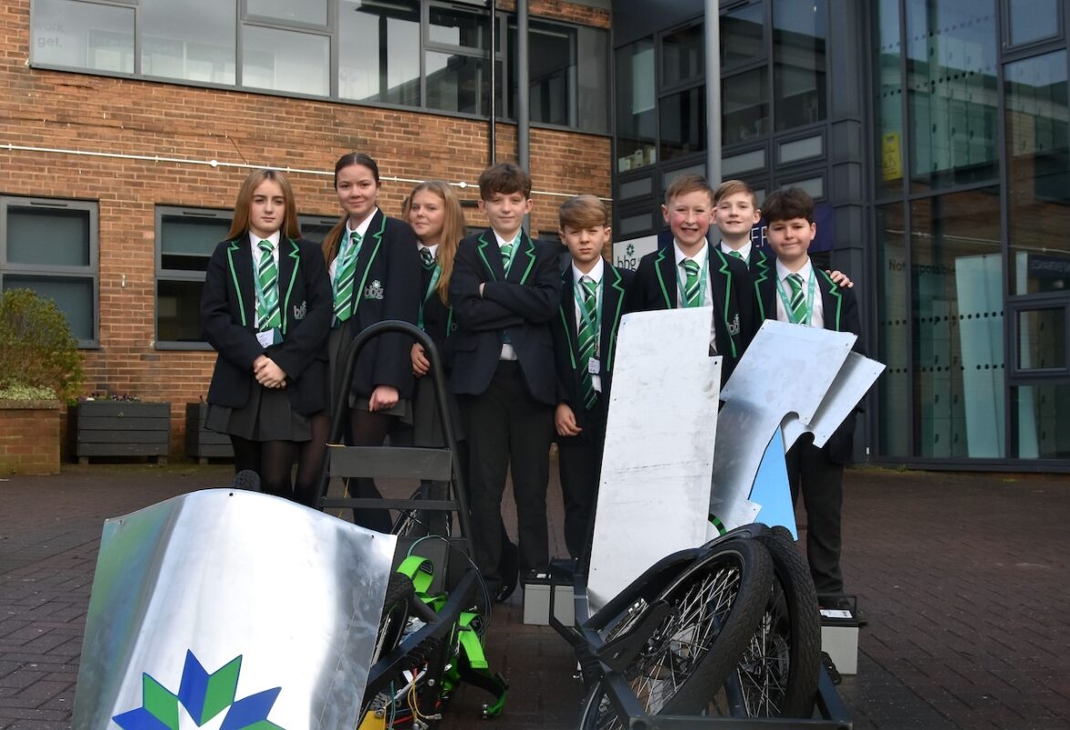 A mixed group of pupils in their school uniforms from BBG Academy in Bradford stand outside their school building behind a greenpower kit car.