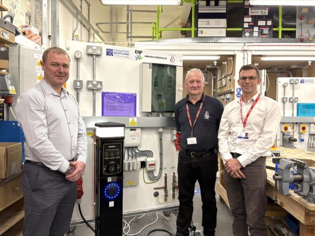 Three people standing in front of an EV Rig within an electrical engineering workshop.
