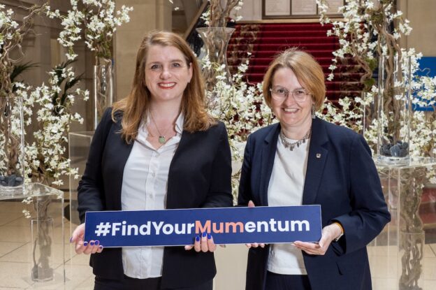 MP Alison McGovern (left) and Professor Josie Fraser from the Open University (right) are standing indoors, holding a rectangular sign that reads "#FindYourMumentum." The background features elegant decor with white floral arrangements and a red-carpeted staircase.