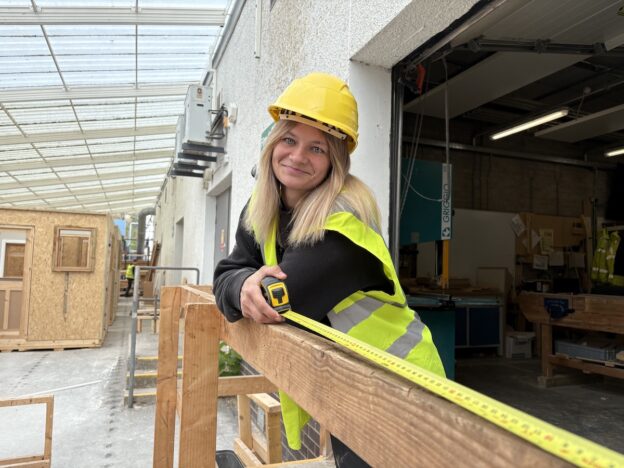 Photo of a female carpentry student standing holding a tape measure in a carpentry workshop